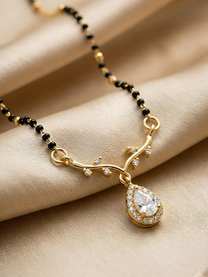 Gold and black beaded necklace with a teardrop-shaped pendant set with clear stones, displayed on a beige satin fabric background.