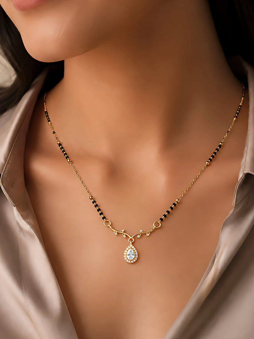 A close-up of a woman wearing a delicate gold necklace with small black beads and a teardrop-shaped clear stone pendant, styled with a champagne satin shirt.