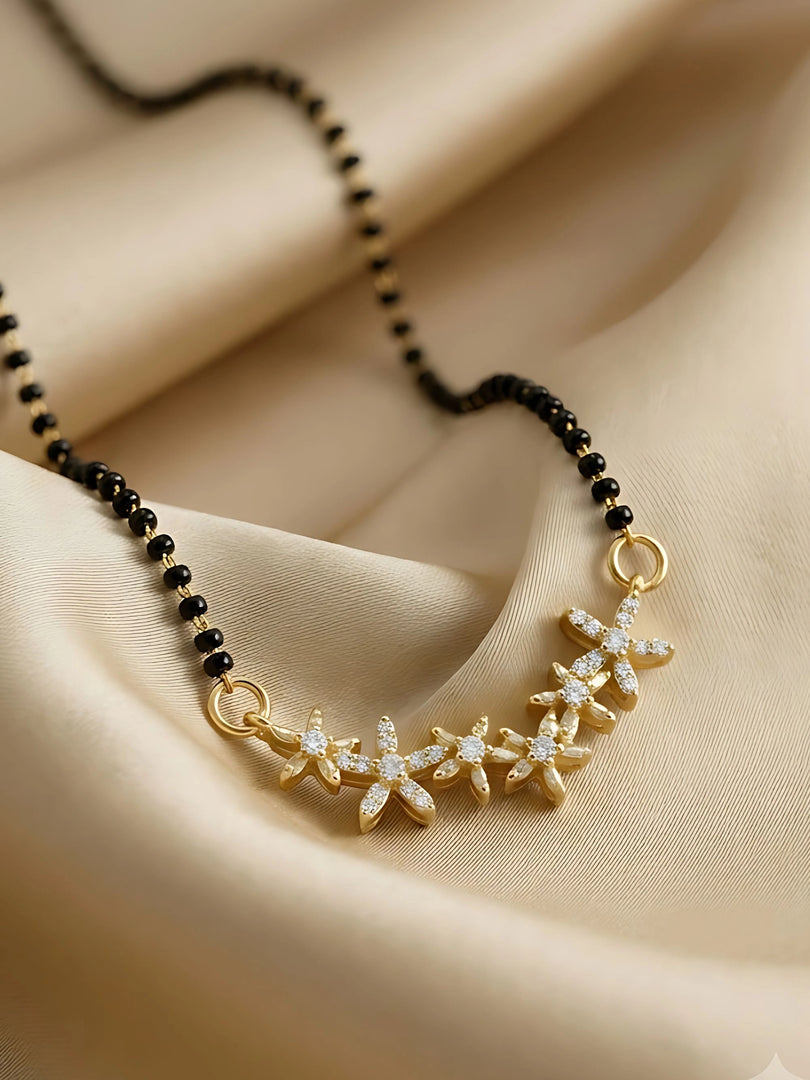 A gold-toned floral necklace with clear rhinestone accents on a black beaded chain, displayed on draped beige satin fabric.